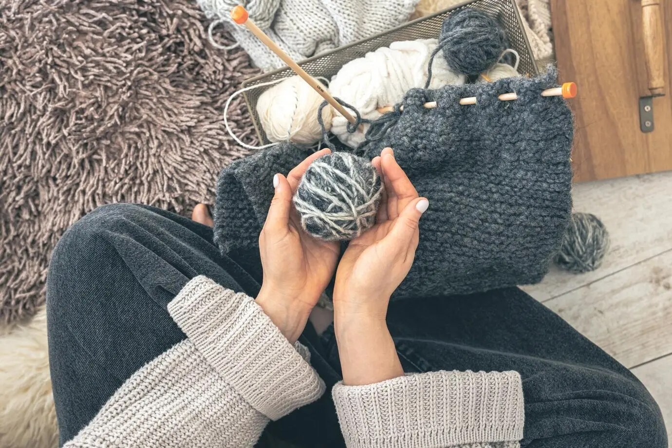 Eine Frau strickt mit Garn, während sie zu Hause auf dem Boden sitzt, in Draufsicht.